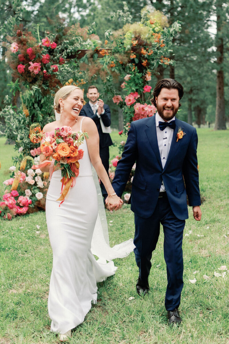 Kelly and Joe standing in front of their floral wedding arbor with coral, peach, and blush peonies at their outdoor ceremony in Bend, Oregon, designed by Summer Robbins Flowers