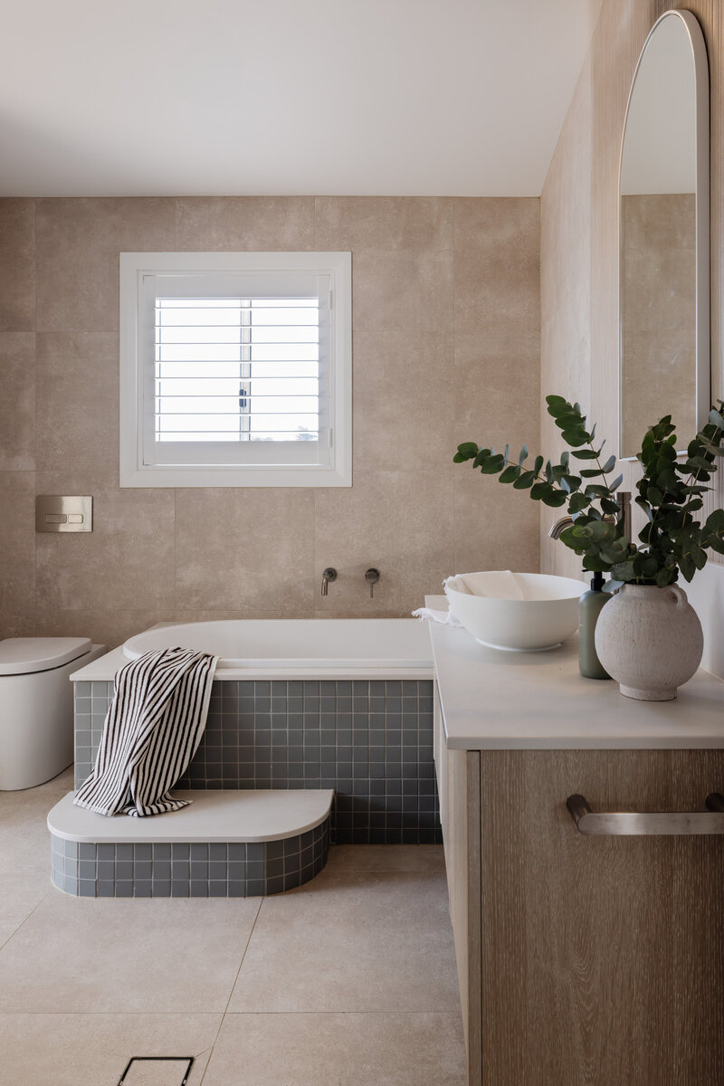 Modern bathroom featuring a bathtub, potted plants, and a round mirror. The window with white shutters adds elegance, crafted by Newcastle's full-service interior design experts for a personalized touch.