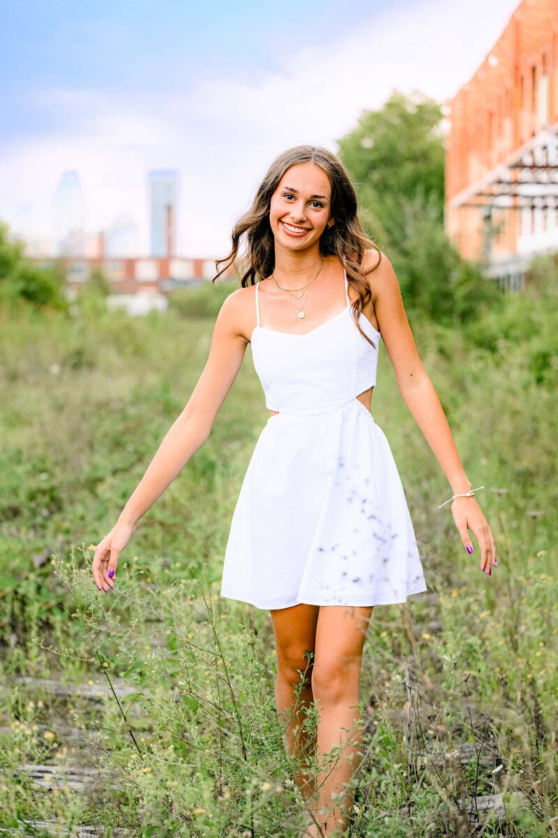 senior in high school wearing white dress  with charlotte skyline behind her