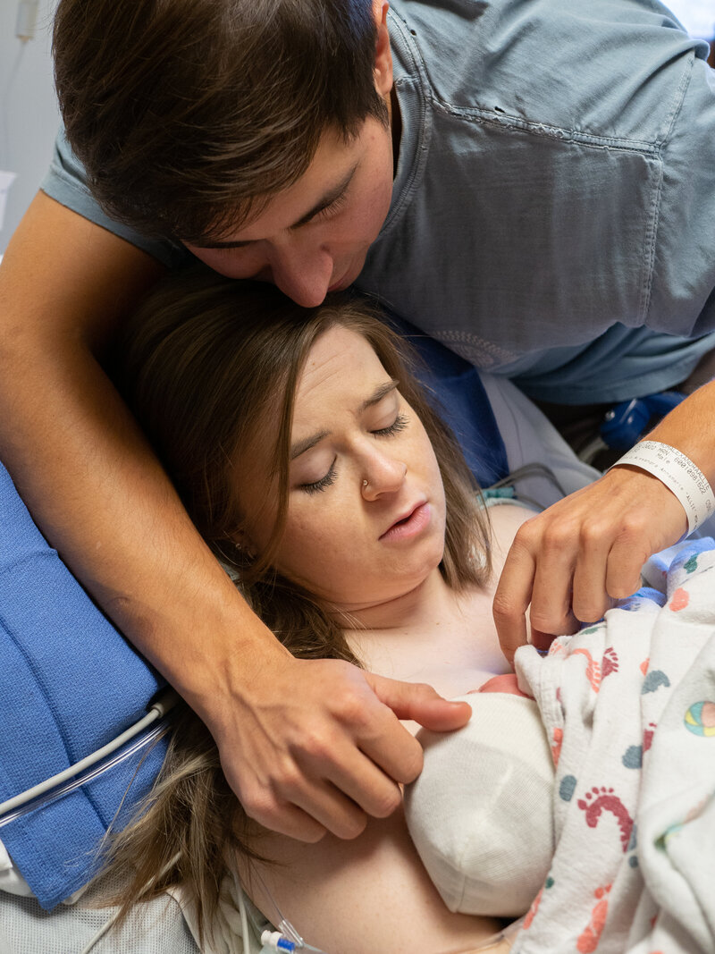 A woman has just given birth and is holding her new baby as she lays in her hospital bed. Her partner is leaning over her and tenderly touching the baby's head.