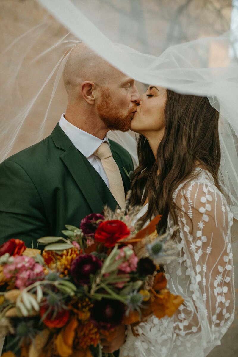 A bride and groom share a kiss under a sheer veil. The groom wears a dark green suit and gold tie; the bride wears a lace dress and holds a bouquet of flowers, capturing an elegant moment perfect for showcasing on your showit web design for wedding planners.