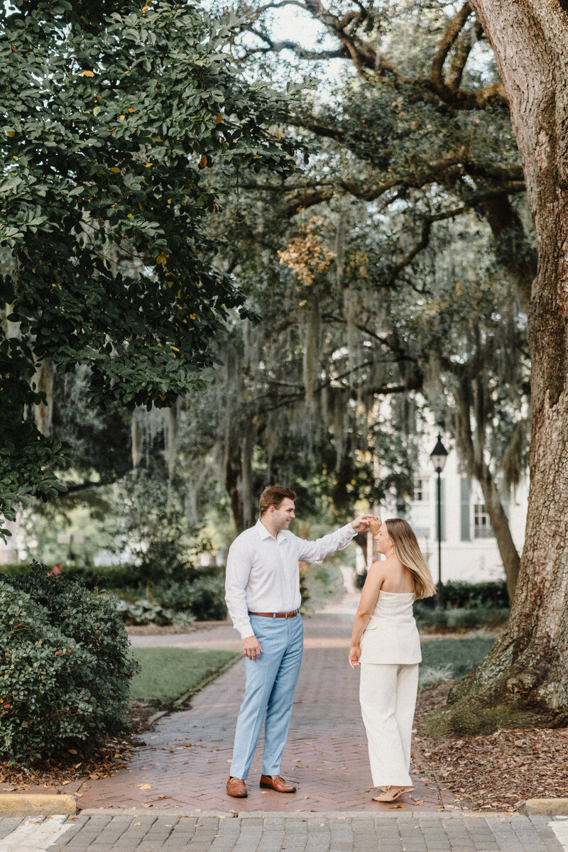Groom in white button up and baby blue slacks twirls bride in creamy white pant suit under historic savannah oaks draped with spanish boss. There are white bricked historic homes in the background.