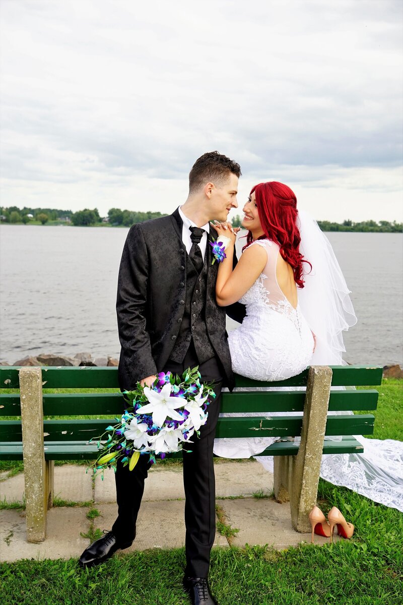 A wedding couple shares a romantic moment in a park, with the groom leaning against the back of a bench and the bride sitting on it, both looking into each other's eyes. This intimate image beautifully illustrates the genuine connection and love we capture in our wedding photography, showcasing our commitment to creating heartfelt and memorable moments.
