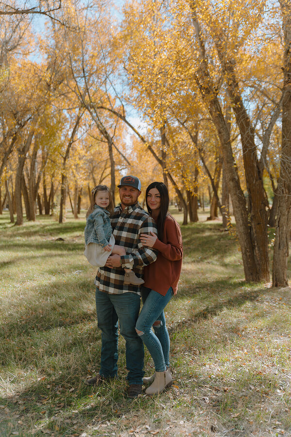 A dad holds his young daughter as his wife holds his arm. Surrounding them are trees with yellow leaves.