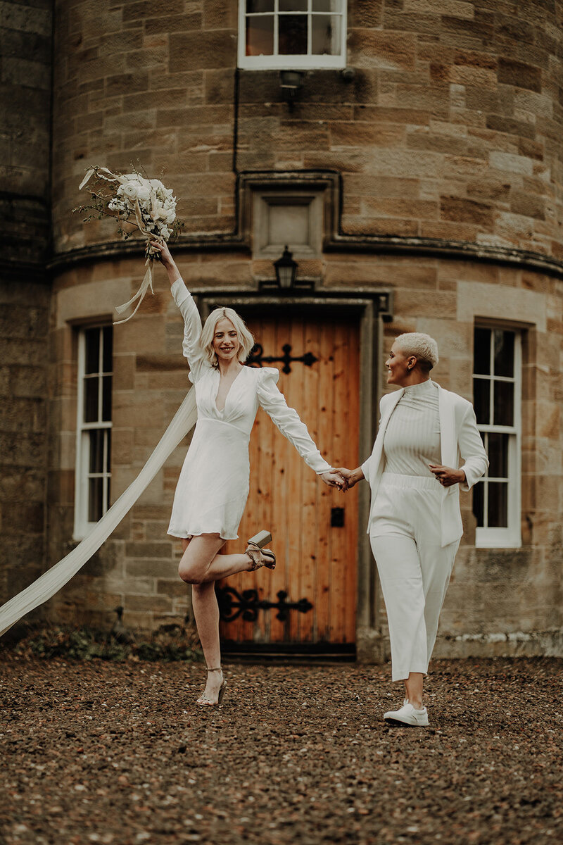 two brides in white outfits holding hands one bride holding her bouquet above her head with round stone door entrance in background fun aberdeen wedding photography
