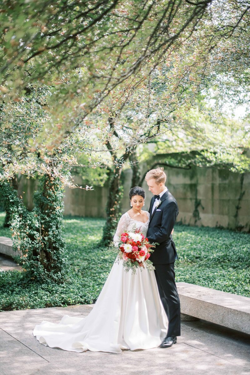 Bride and groom standing beneath a canopy of trees in a quiet garden, bride holding a bouquet of red and white flowers while groom embraces her.