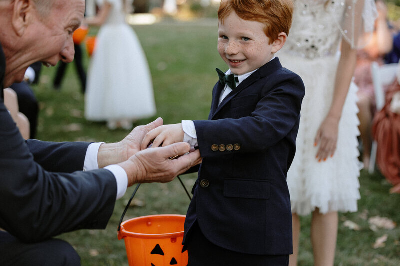 A young ring bearer in a suit offers halloween candy to guests. He carries an orange Halloween bucket 