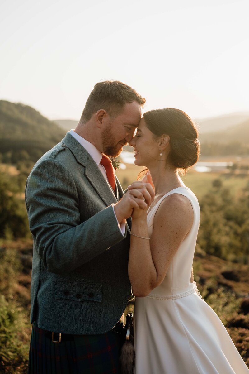 A couple stand with their chests together and arms extended. The link fingers and rub roses romantically. The wide photography shows golden hour on the Mar Lodge Estate and a large golden valley can be seen behind the couple. They wear wedding attire and the sun flickers between them.