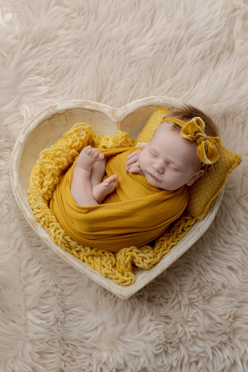 Lehigh Valley Newborn Photographer LG Photography Lori Generose photo of a newborn baby girl in mustard yellow wrap and on a blanket in a cream wooden heart bowl on a cream colored rug