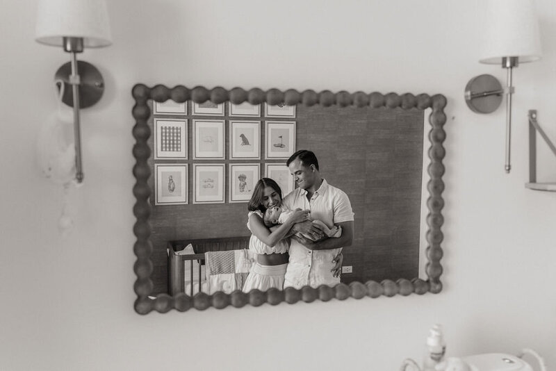 Black and white photo of a couple holding their newborn, reflected in a mirror above a changing table. The reflection shows the family standing in a softly lit nursery with framed animal prints on the wall behind them.