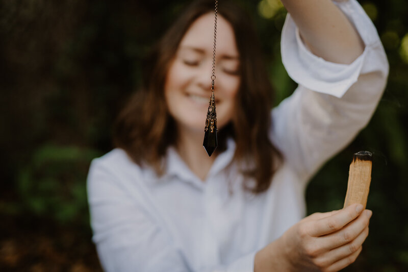 Brandi of the Intuitive Momma smiles with her eyes closed and holds a pendulum and Palo Santo