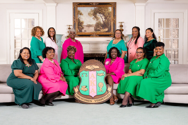 PAO Chapter offiers wearing pink and green, sittign in a formal living room behind a giant wooden, AKA  shield