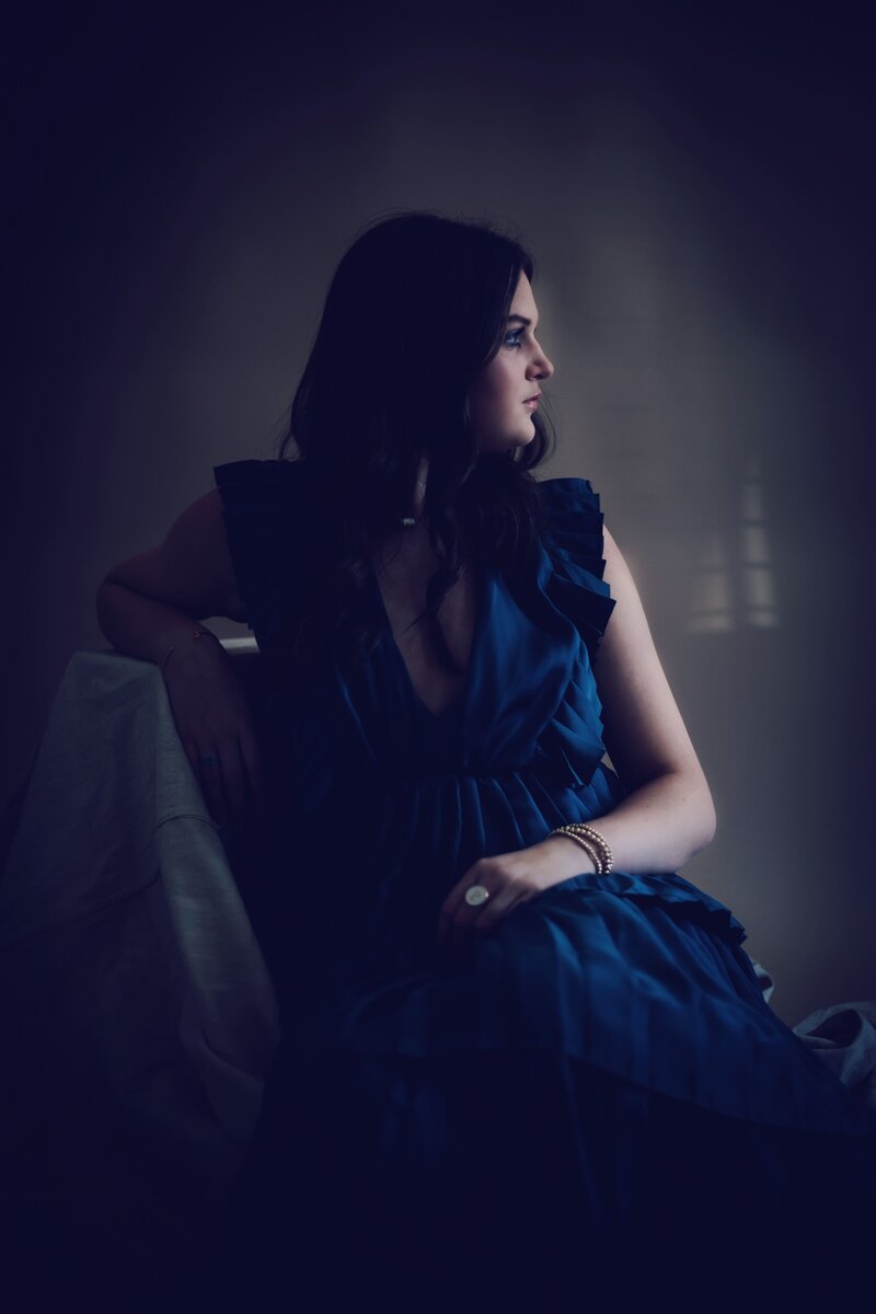 A woman in a blue dress sitting in a dark room, captured by a talented senior portrait photographer Britt Elizabeth.