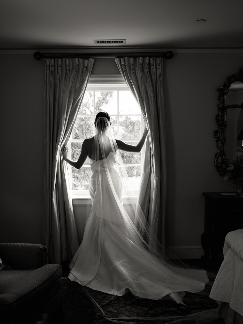Julia stands by the window at Boxwood Estate, gazing out as she prepares for her wedding day. She wears a beautiful wedding dress with intricate details and a flowing veil. The soft light filters through the window, creating a serene and intimate atmosphere. Her calm expression reflects the anticipation of the special day ahead, with the elegant interior of the estate adding to the moment’s beauty.