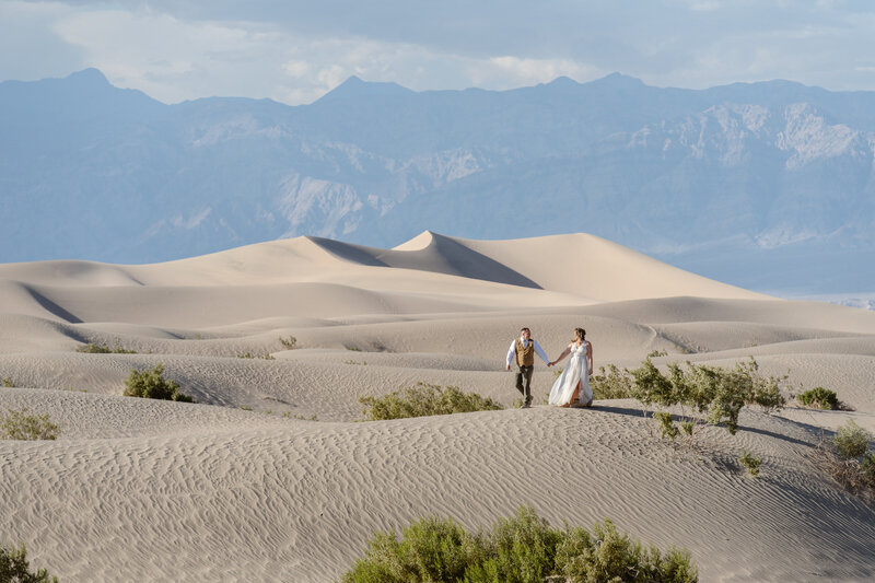 A couple in wedding clothes walks hand in hand along sand dunes