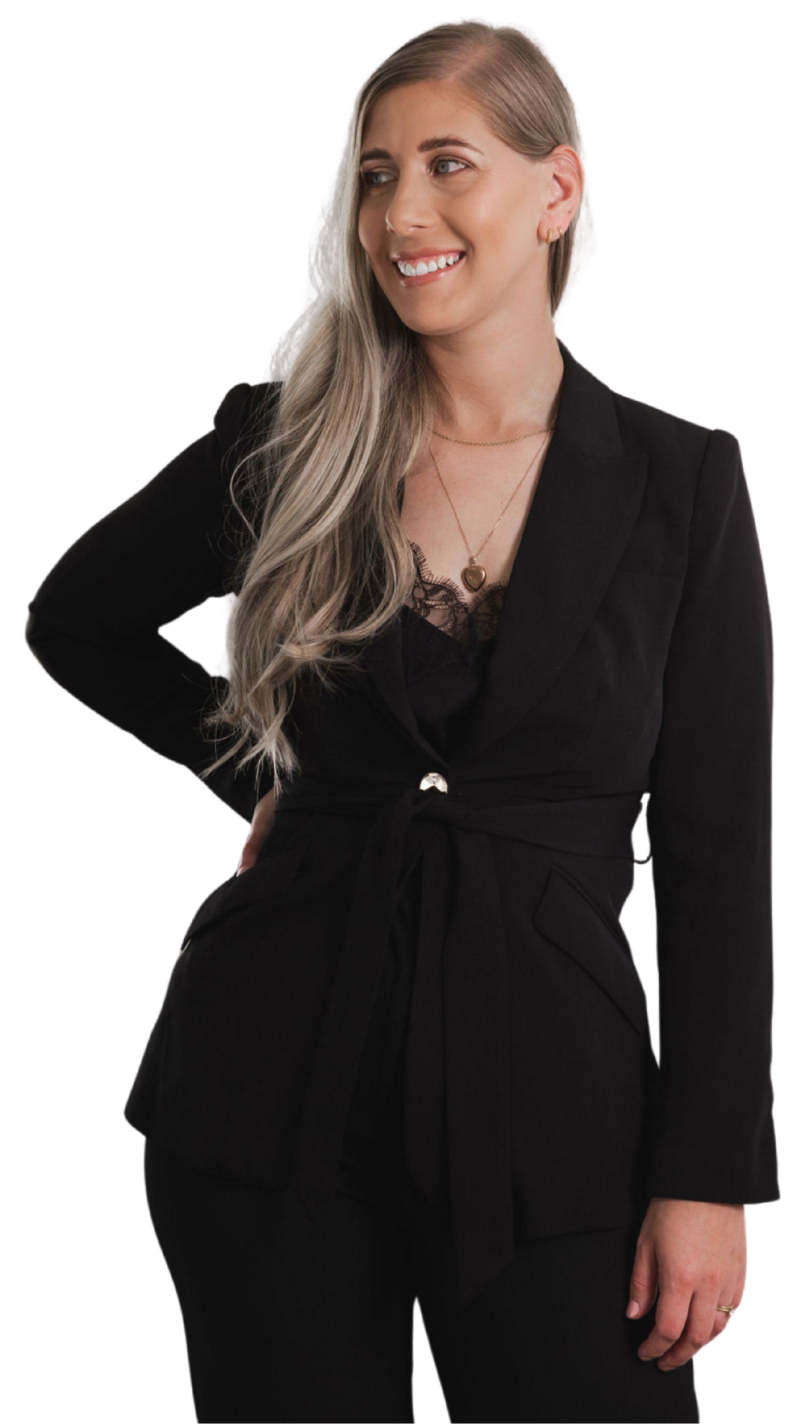 Woman smiling while wearing a black blazer with a belted waist and lace top, standing confidently with one hand on her hip.