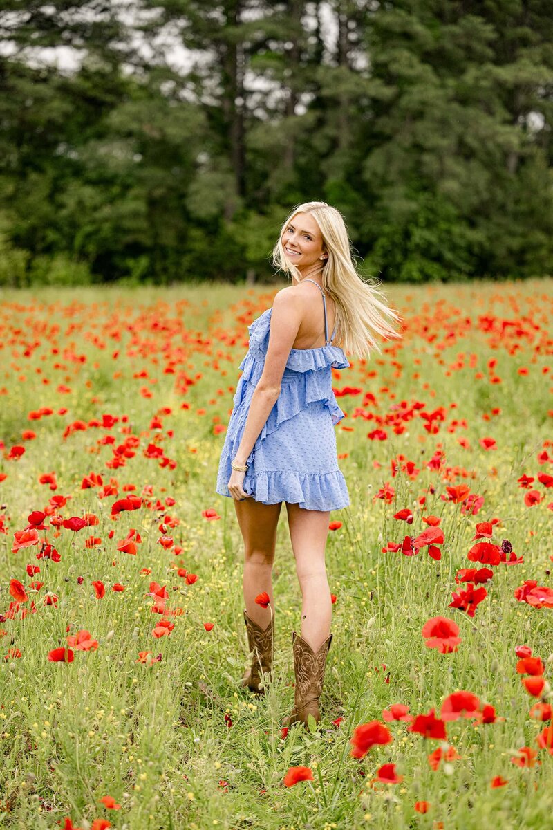 blonde charlotte graduate walks in red poppy field wearing a blue dress and brown boots for her senior photos