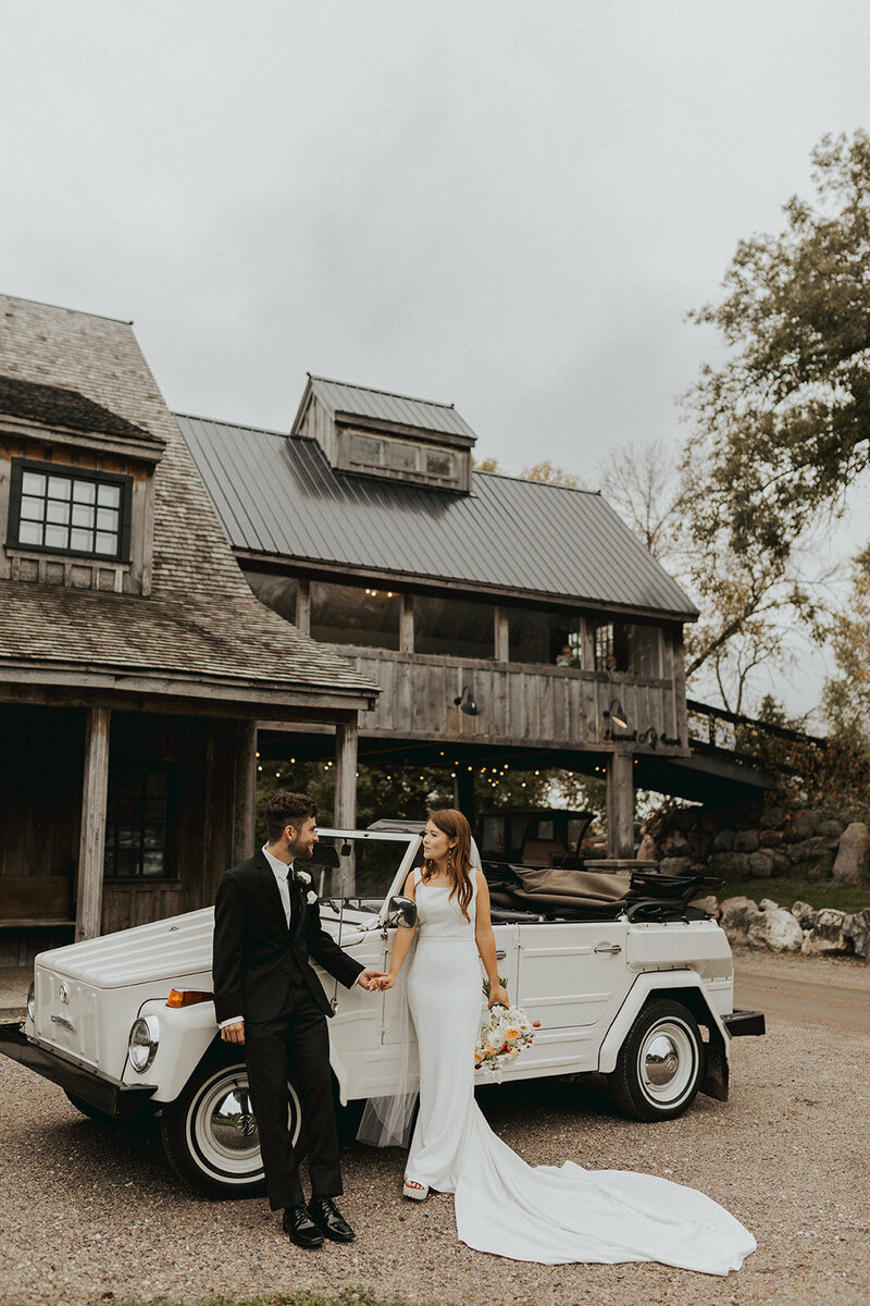 Bride and groom standing hand in hand beside a retro car, gazing affectionately at each other – a timeless moment captured in love.