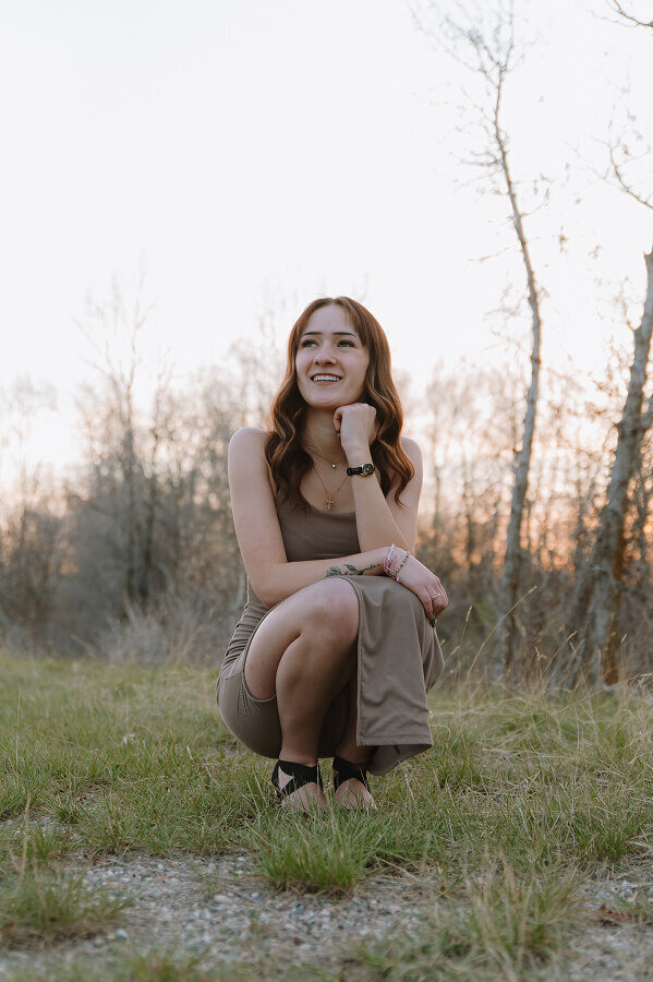 A high school senior graduate smiles for her senior photos. She is kneeling close to the ground and looking off into the distance.