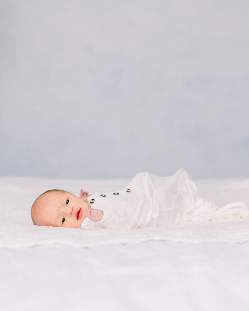 Newborn baby in white knotted onesie, laying on white bedding in front of a blue and white hand painted backdrop, by Boston family photographer Fieldstone Studio.