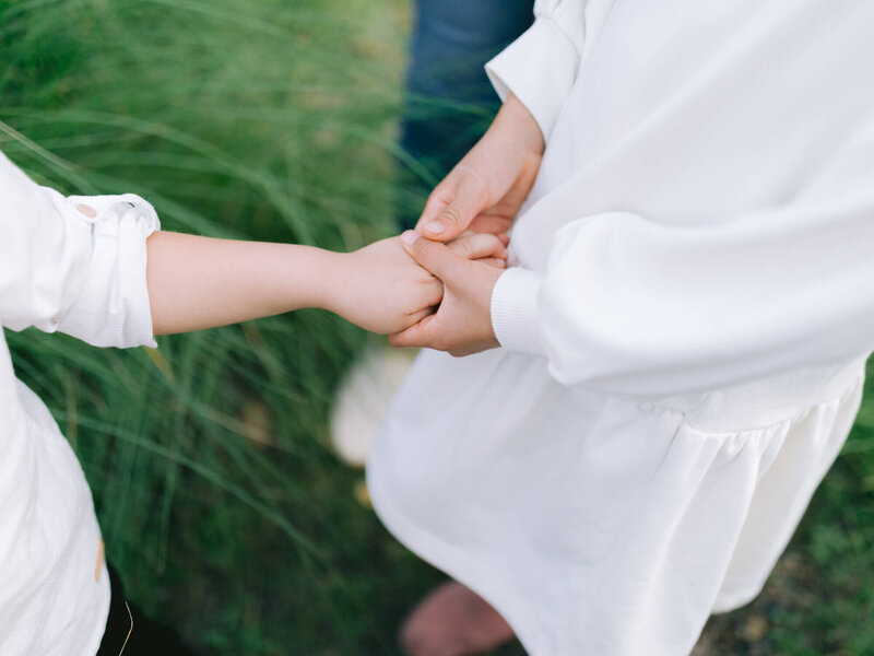 Two kids in white clothing gently hold hands against a blurred background of green grass, conveying a sense of connection and tenderness.