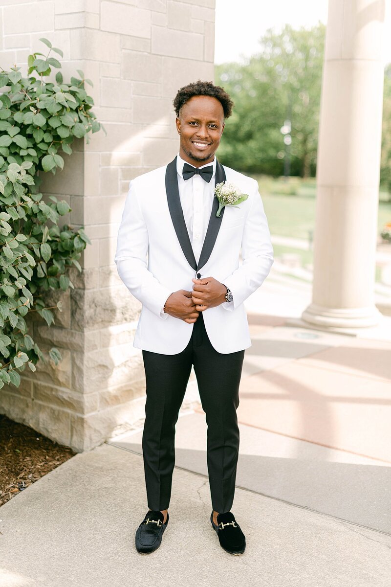 A groom wearing a white tuxedo jacket and black pants stands on the side walk at Coxhall Garden. He is smiling at the camera and has a bowtie and  velvet shoes
