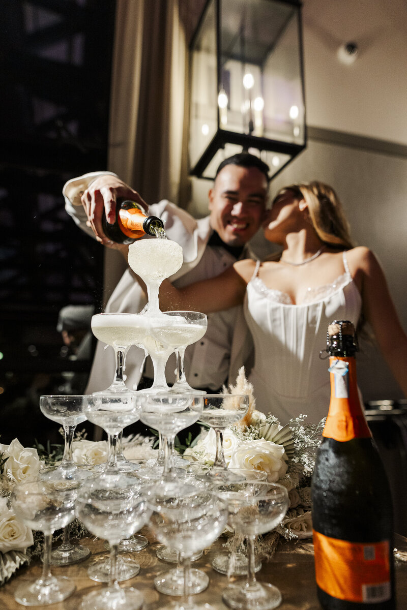 Evert pouring a champagne tower while Haley kisses his cheek at their wedding reception