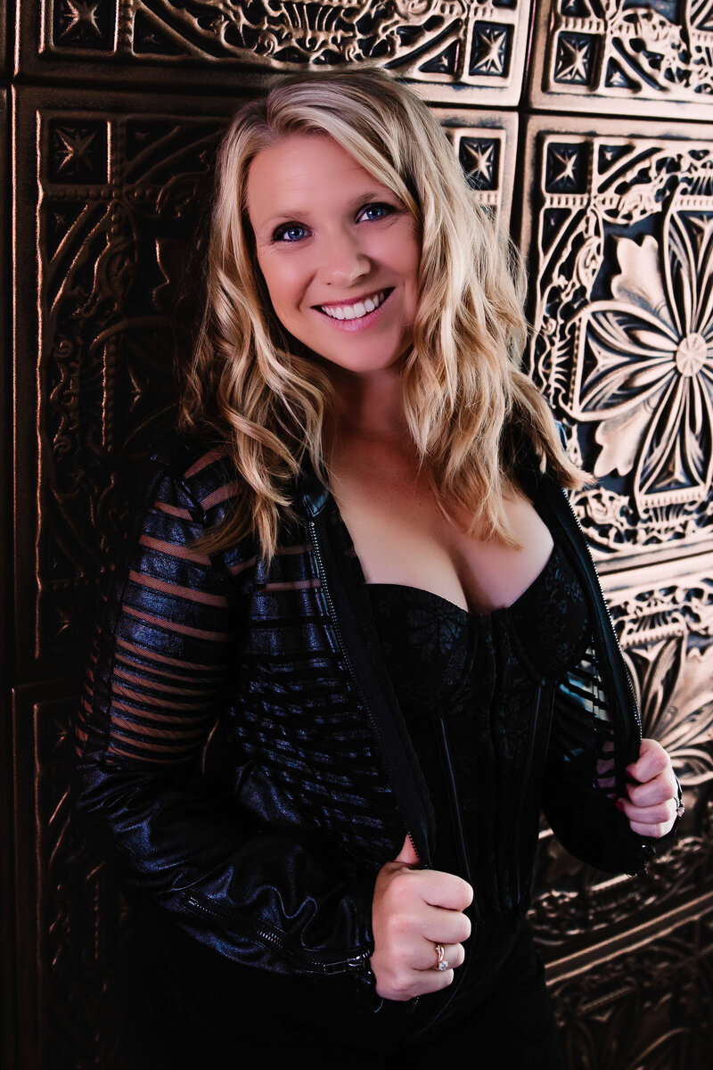blonde woman leans against gold wall in leather jacket and bustier