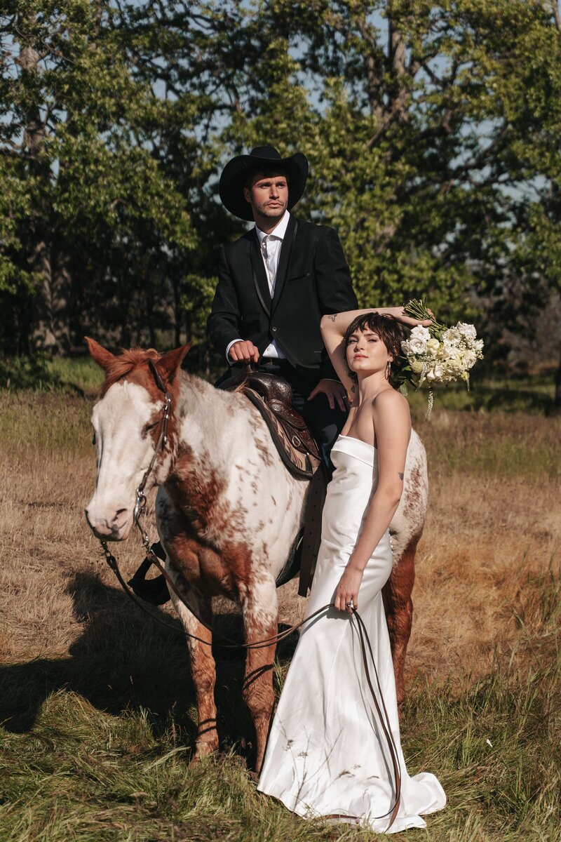 Bride and groom in wedding attire pose editorial-style with a horse on a sunlit hillside in the Columbia River Gorge, holding a white bouquet on a spring day.