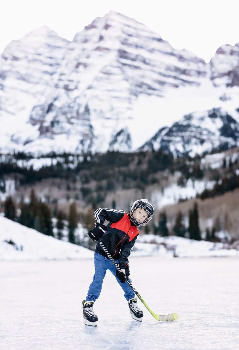 A little boy posing with his hockey stick in front of the Maroon Bells, skating on a frozen Maroon lake.