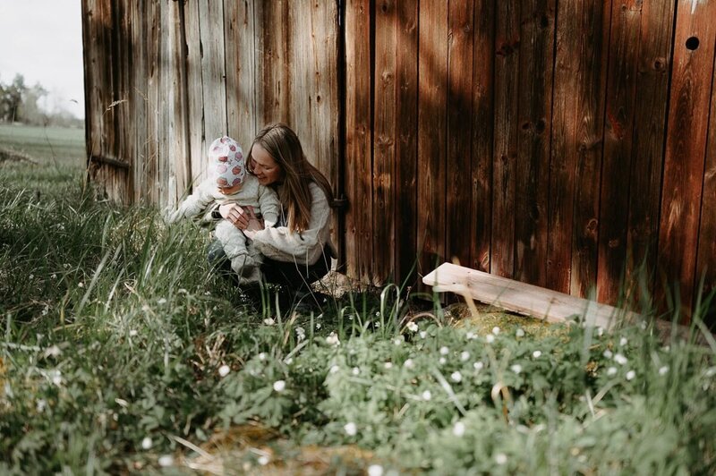 Mamma och dotter sitter i gräset på huk framför en ladugårdsvägg.