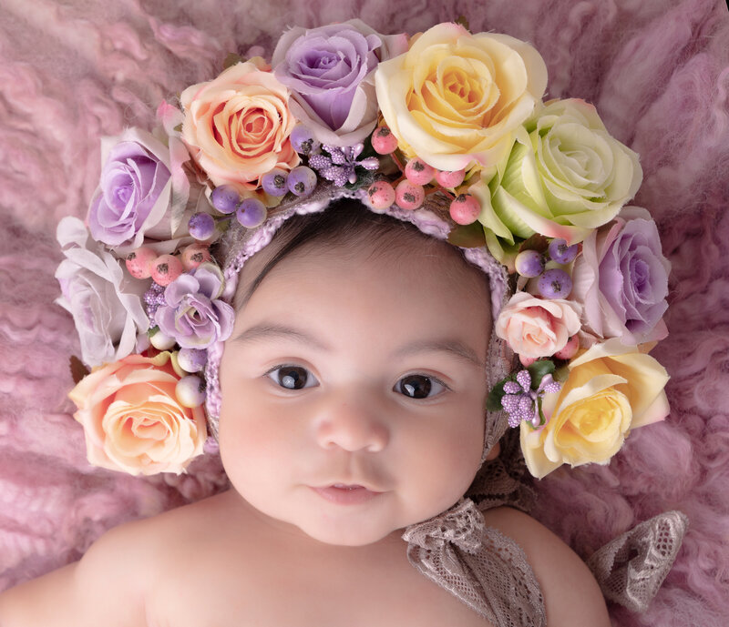 A baby with a serene expression wears a vibrant flower crown of roses in purple, pink, and yellow. The soft pink blanket adds a cozy, gentle backdrop.