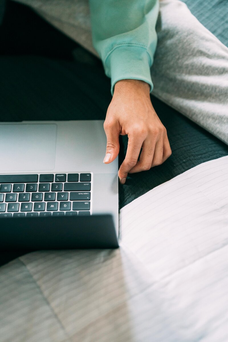 Close-up of a person in a green sweater using a laptop on a bed, preparing for an online lesson


