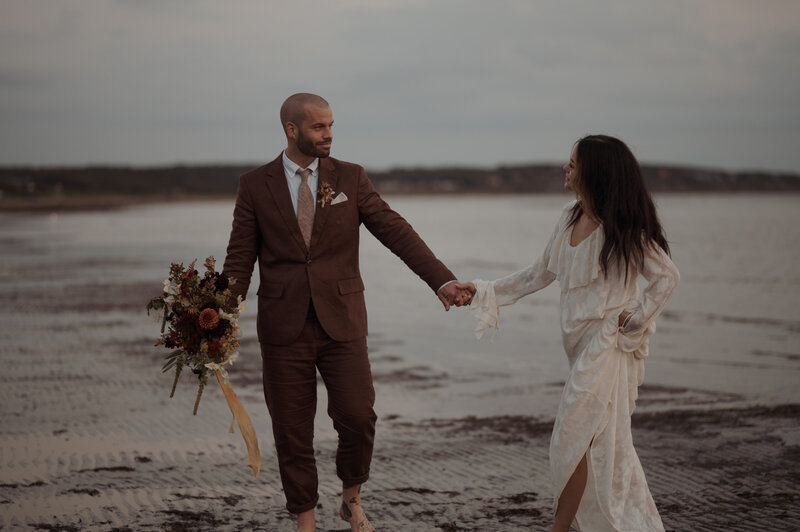 Brudpar går längs havet och håller varandras händer i solnedgången.