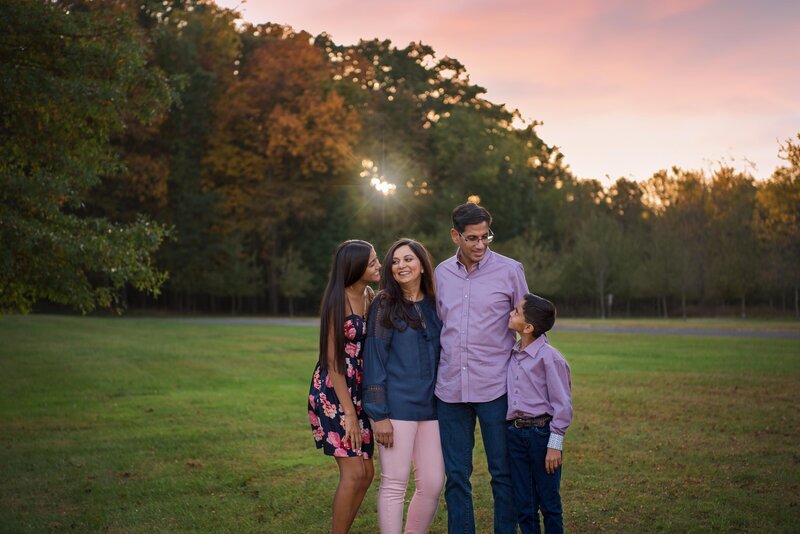 A family of 4 looking at each other and smiling for their family photoshoot at a park in New Jersey during sunset.