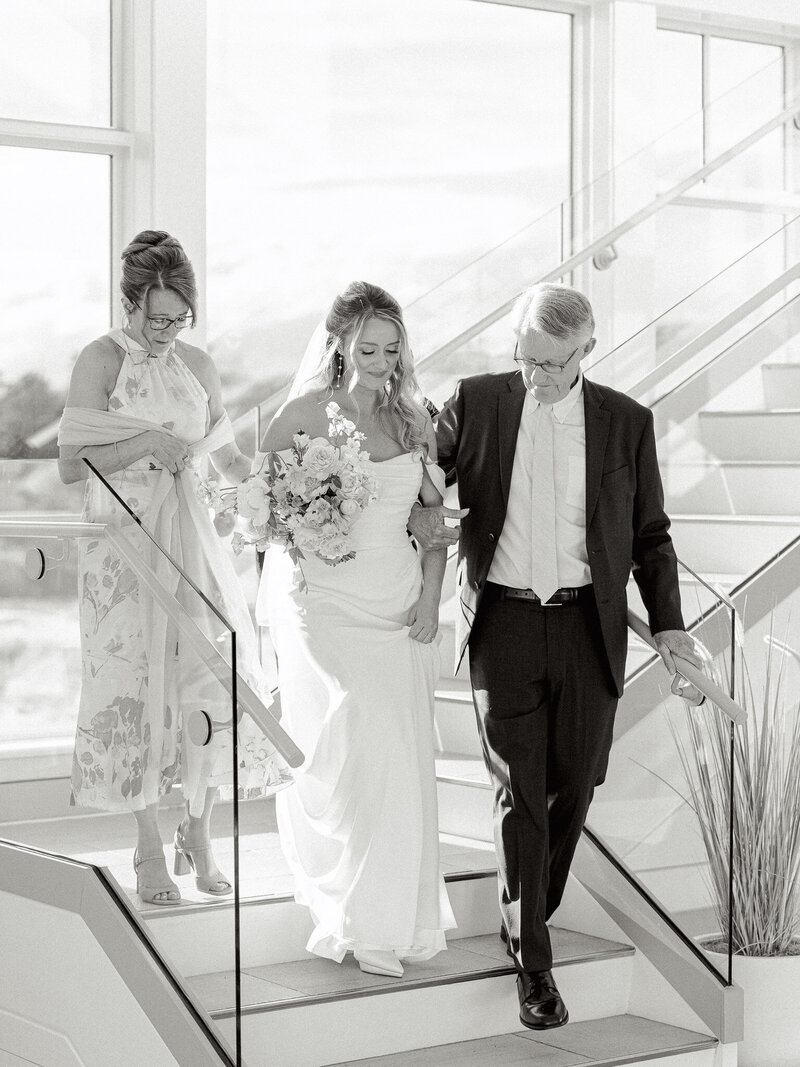 Destination black and white wedding photo of the bride and her parents walking down the stairs towards the ceremony.
