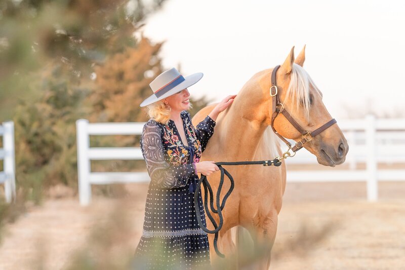 Dressed for fall or winter, a blonde woman in a grey felt western fashion hat pets her palomino reining Quarter Horse at her private stable in Edmond Oklahoma