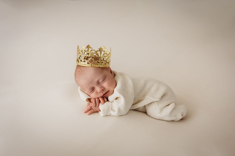 Serene studio portrait of a newborn baby sleeping soundly on a plain light beige surface, adorned with a beautiful gold crown and wearing a soft white romper