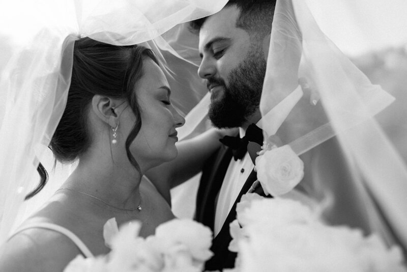 Emotive black and white portrait of bride and groom under wedding veil.