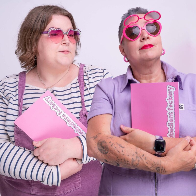 Two people pose side by side against a pale studio backdrop, each hugging a bright pink notebook. Left is Lucy in a striped top with purple overalls and rose-tinted sunglasses. Right is Pippa in lilac dungarees with round pink heart-shaped sunglasses and tattooed forearms, both striking a playful, mock-serious stance.