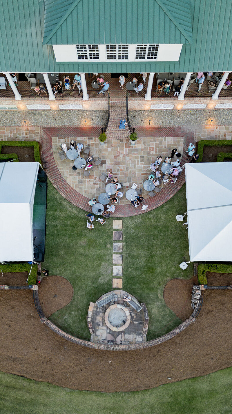 Aerial view of an outdoor event at Woodcreek Country Club in Columbia, SC, showing guests gathered around round tables on a stone patio. A grand staircase connects the covered veranda to the landscaped lawn, where two white tents frame the event space. A stone firepit sits at the center, adding to the elegant atmosphere. Small business branding photography in Columbia, SC, highlighting luxury event venues and corporate gatherings.