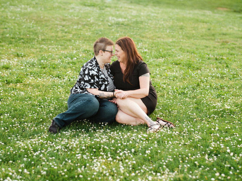 LGBTQ couple sit on the grass and laugh during an engagement shoot
