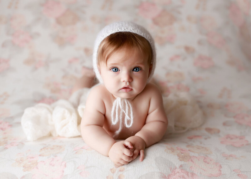 Studio portrait by best newborn photographer in West Palm Beach Florida. baby is laying on a floral fabric, propped up on arms wearing a white knit bonnet. Baby girl has red hair and blue eyes and looking straight at the camera.