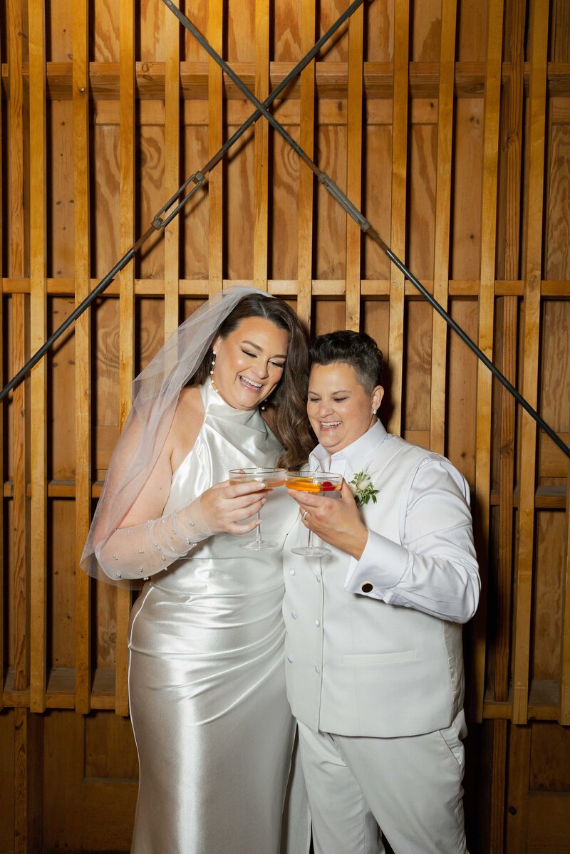 Two brides smile and clink cocktail glasses.
