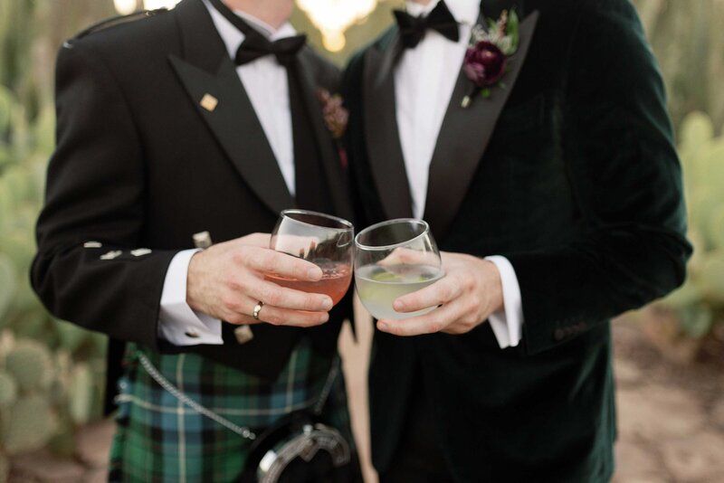Two grooms clink cocktail glasses during sunset photos.