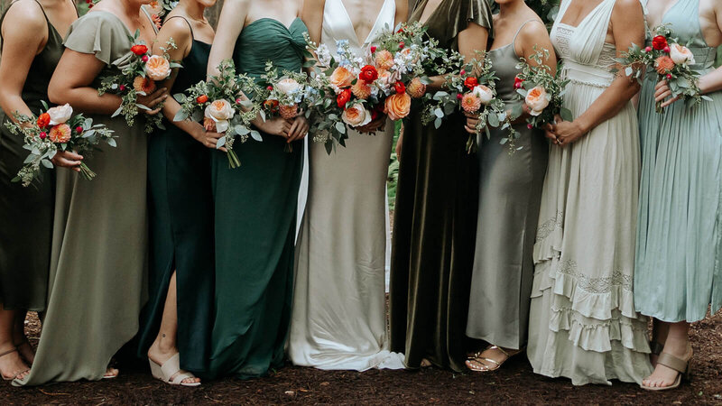 Bridesmaids with dresses in shades of green and carrying bright bouquets huddle together.