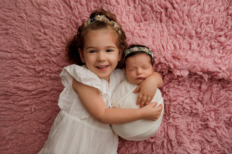 A mother gently cradles her newborn baby against her chest, both looking peaceful and content. The mother has long, straight black hair and is wearing a soft, off-the-shoulder white sweater. The newborn is swaddled in a diaper and has a delicate headband with a floral design. The background is a warm, light brown, adding a cozy and intimate feel to the portrait. The mother's serene expression and the baby's relaxed pose convey a sense of love and tenderness.