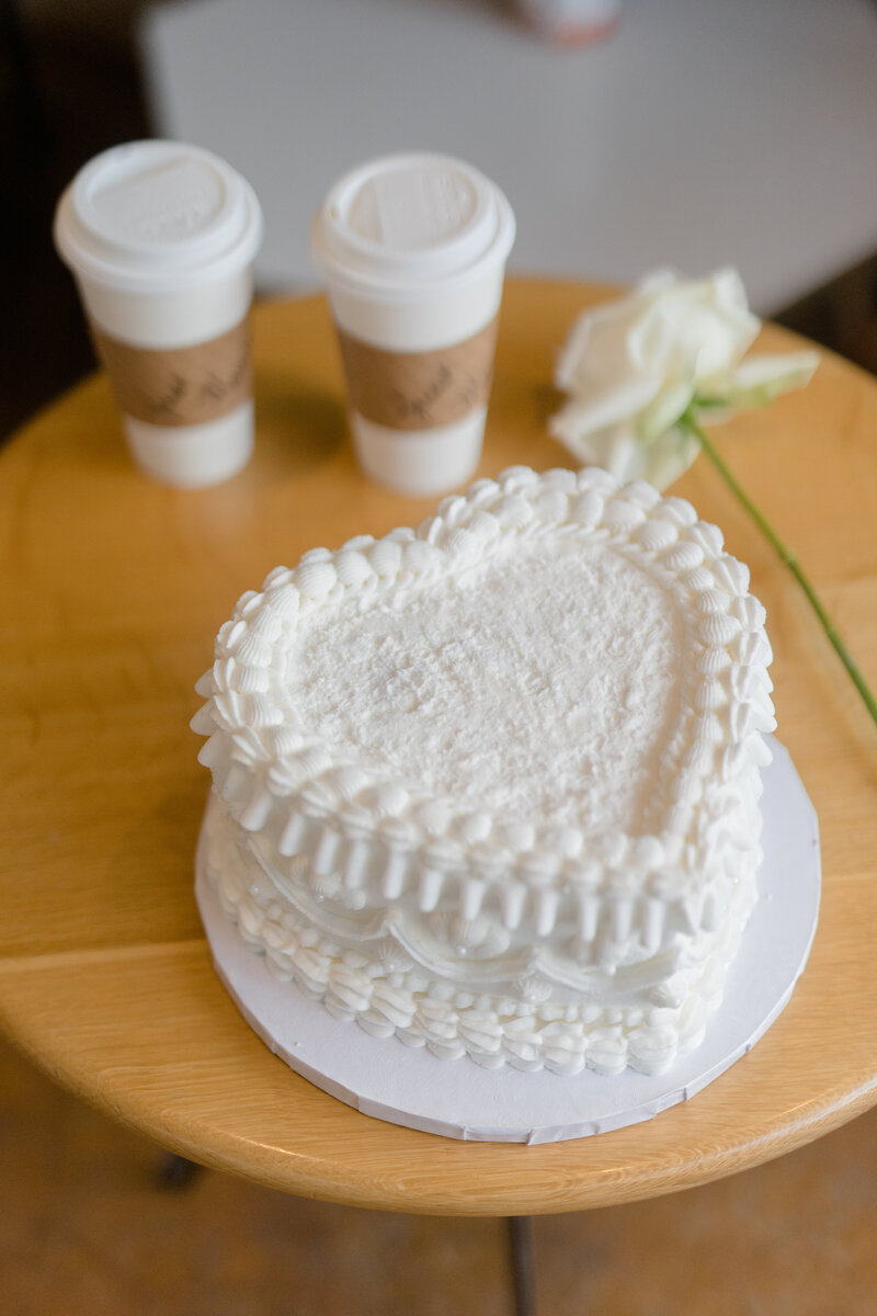 Aerial view of a white heart shaped one-tier vintage piped wedding cake sitting on a low table with two togo coffee cups in the background and a single white rose at Compass Coffee in Georgetown DC 