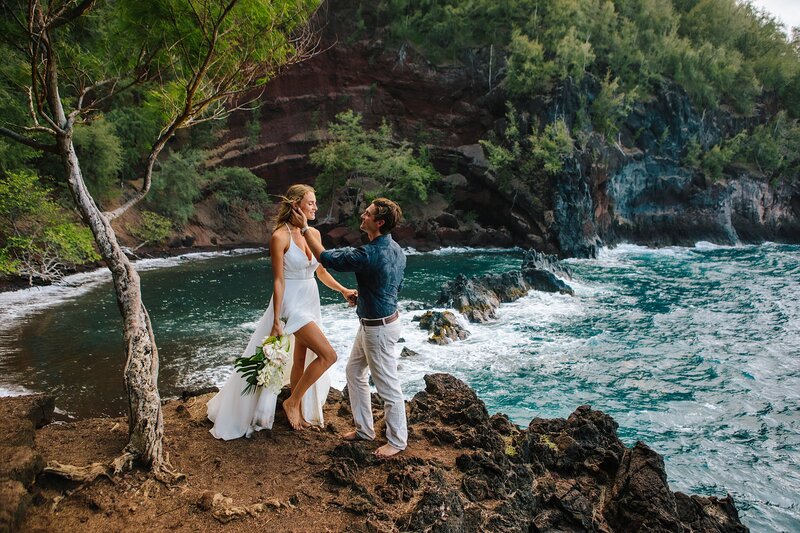 A bride and groom stand on a cliffside overlooking a beach with red sand in Hawaii