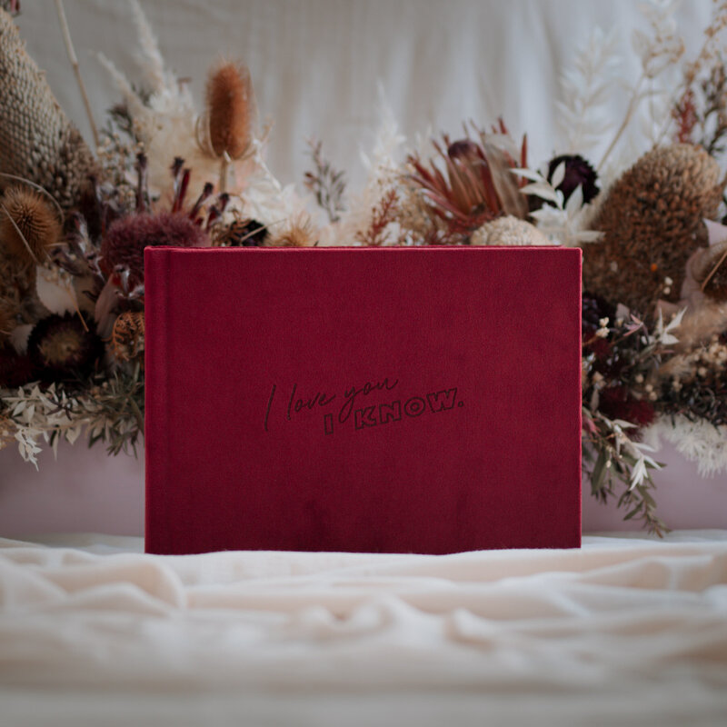 Burgundy velvet guest book with custom engraving on the cover in front of a dried flower arrangement